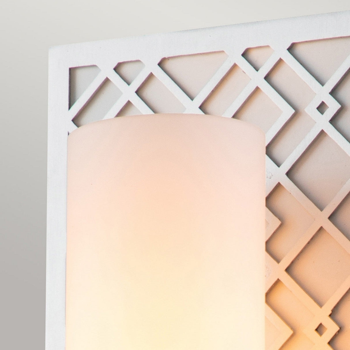 Close-up of the Ziggy 1 Light Wall Light - Lacquered Silver, showcasing its opal glass shade against a decorative geometric lattice backdrop. The fixture's frosted design delivers a soft, warm glow, creating gentle shadows on the white patterned surface and enhancing its lacquered silver finish.