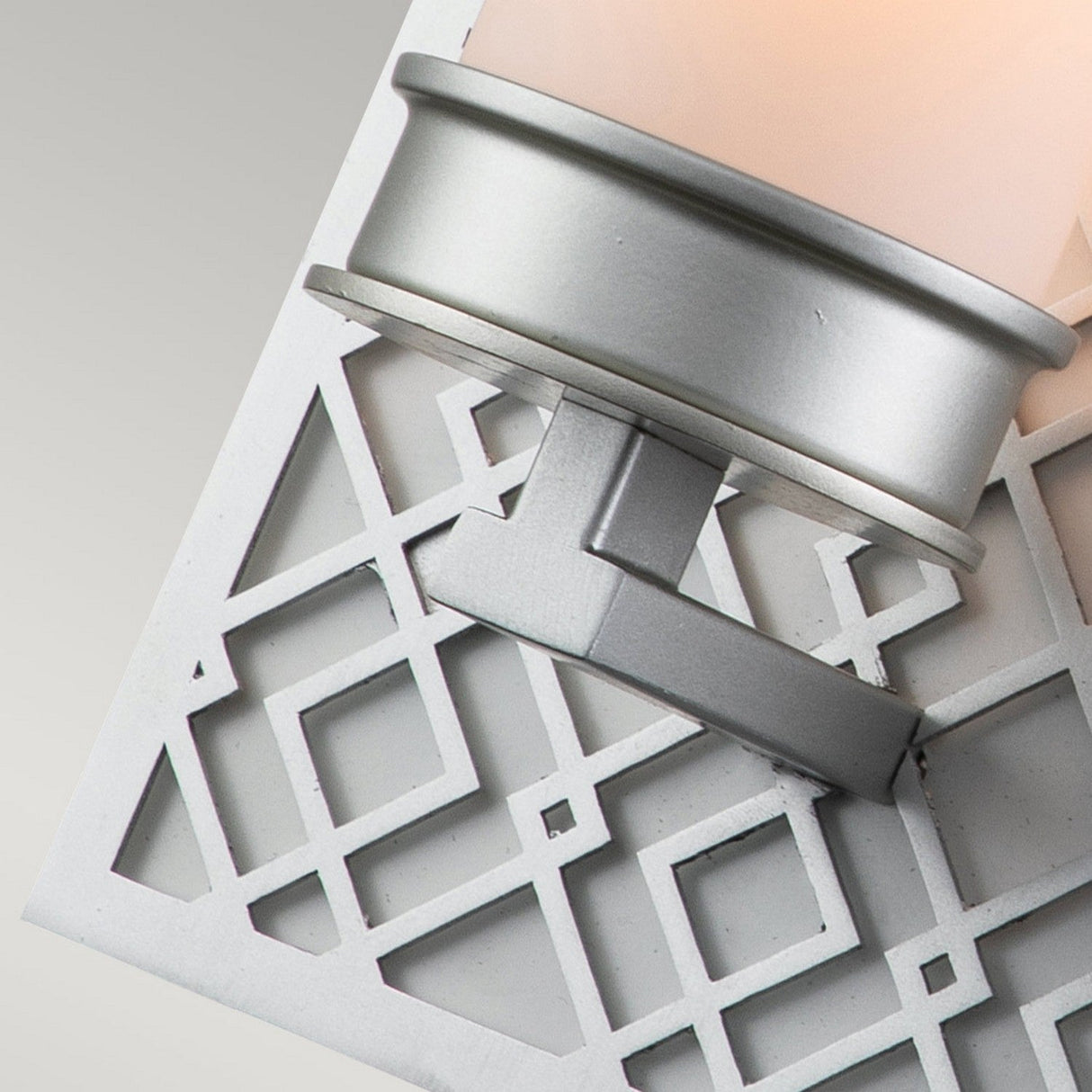Close-up of the Ziggy 1 Light Wall Light in Lacquered Silver, a contemporary sconce boasting a geometric design. It showcases intersecting diagonal lines on its finish and features a cylindrical opal glass shade against a lattice-like background, softly illuminating the area.