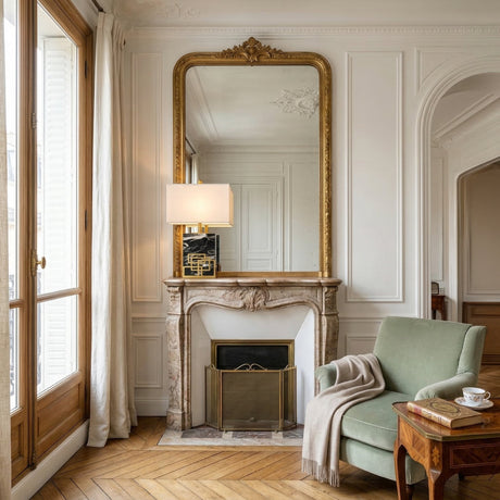 Elegant living room with a marble fireplace, gold-framed mirror, Veridan 1 Light Framed Marble Table Lamp in Brass & Black, pale green armchair with blanket, side table with tea set, herringbone wood floors, and tall window with white curtains.