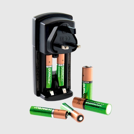 A black battery charger is plugged into a wall, charging four Duracell rechargeable batteries. Four additional batteries are scattered below it, two with golden tops and two green cylindrical ones.