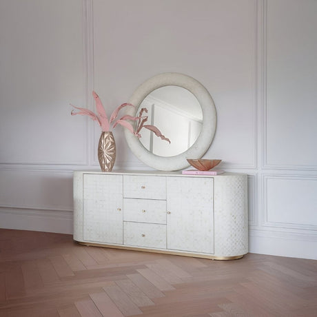 White textured sideboard with a large round mirror, pink floral arrangement, and warm décor set in a bright, elegant room with herringbone floors.