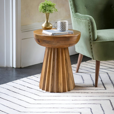 Wood pedestal side table with decorative items beside a green chair, styled in a bright corner with a modern rug for a warm, inviting touch.