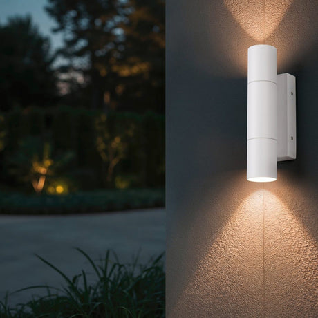 Yamuna 2 Light Up Down Outdoor Wall Light in matt white aluminium, mounted on a textured exterior wall casting warm upward and downward light, with a garden setting visible at dusk.