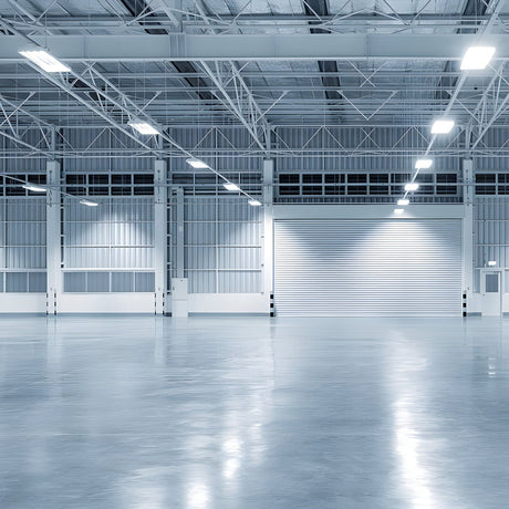 A spacious, empty warehouse with polished concrete floors, high ceilings, and large overhead lights. The walls are lined with metal panels, and a large closed roller door is visible at the back.