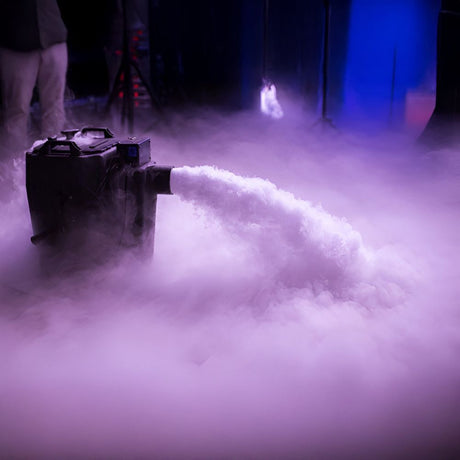A fog machine producing a thick stream of white fog across a dimly lit stage floor.