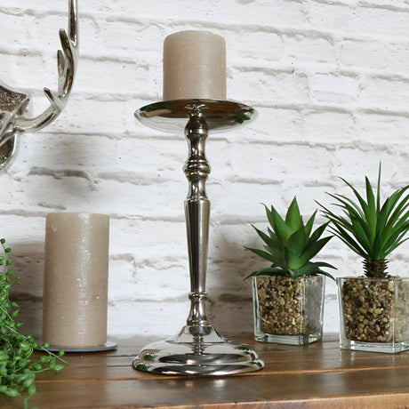 Decorative arrangement featuring a tall silver candle holder, small plants, and neutral candles displayed on a wooden surface against a textured white brick wall for a rustic look.