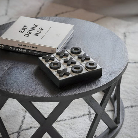 Round black coffee table with an open crisscross base, styled with stacked books and a tic-tac-toe game set on top, placed over a soft patterned rug in a modern living room.