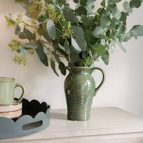 Green ceramic pitcher filled with eucalyptus stems, placed on a light wooden surface beside a small tray, creating a fresh and natural decorative arrangement for the home.
