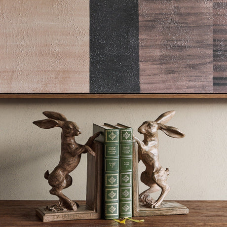 Libra Interiors Rustic Weathered Standing Hare Bookends in brown resin, framing green Charles Dickens volumes on a wooden shelf below abstract art.