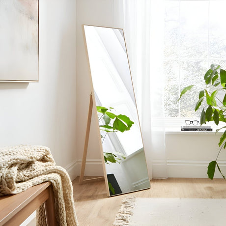 Bright room with a tall, leaning floor mirror reflecting soft natural light. A plant, sheer curtains, and cozy textures create a calm minimalist corner.