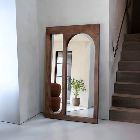 Large arched wooden-frame floor mirror placed near a staircase, reflecting a warm-toned interior with a plant and chair for a rustic, inviting entryway look.