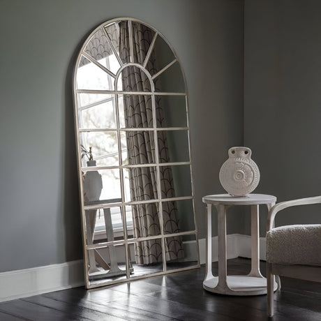 Tall arched mirror with windowpane detailing leaning against a gray wall beside a small white table, adding elegance and height to the room.