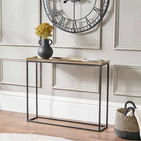 A slim black iron console table with a gold cast top in a panelled hallway, under a large wall clock, with a black vase and a woven basket