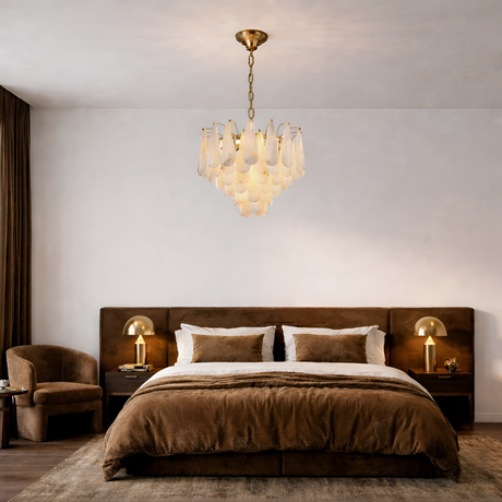 A modern bedroom with a large brown bed, matching brown chair, two nightstands with gold lamps, and a unique glass chandelier hanging from the ceiling.