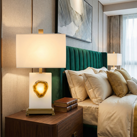 A modern bedroom with a green velvet headboard, beige bedding, and a wooden nightstand featuring a glowing lamp with a stone accent and stacked books. Another lamp is visible in the background.