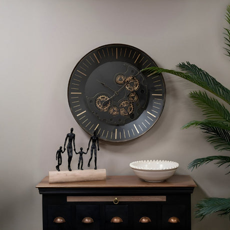 A decorative vignette featuring a large black wall clock with exposed gears, sculptural metal figures, and a textured bowl on a wooden console table.