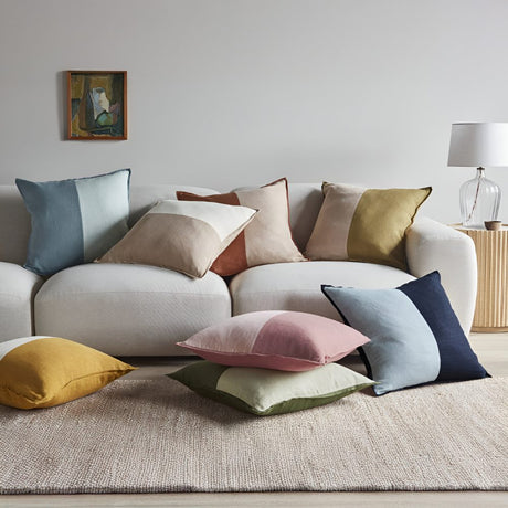 A neutral living room with a white sofa covered in assorted pastel and earth-tone throw pillows, with more cushions scattered on a soft beige rug.