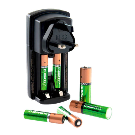 A black battery charger is plugged into a wall, charging four Duracell rechargeable batteries. Four additional batteries are scattered below it, two with golden tops and two green cylindrical ones.