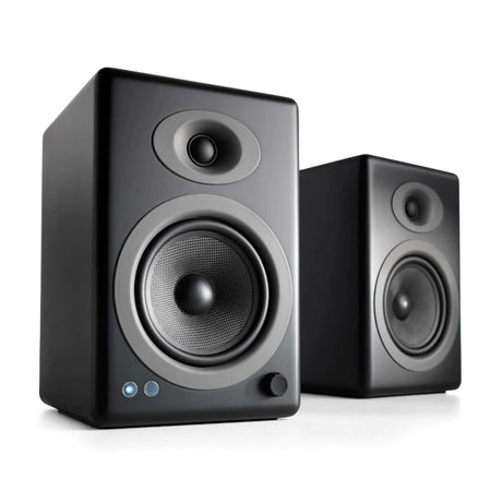 Two black audio speakers with a sleek design. The front speaker features a large woofer below a smaller tweeter. Both speakers are angled slightly, set against a plain white background.