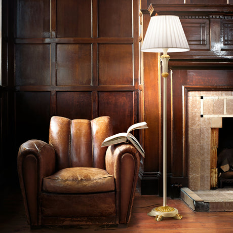 A cozy reading nook features a vintage leather armchair and an open book, illuminated by the Firenze Floor Lamp in White, which combines modern functionality with its pleated shade. Set against dark wood paneling and a tiled fireplace, this arrangement creates warm ambient lighting and an inviting atmosphere.