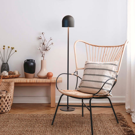 Rio 2 Light Dome Floor Lamp in matt black, slim stem and round base, shown beside a rattan chair in a living room with rug and side table.