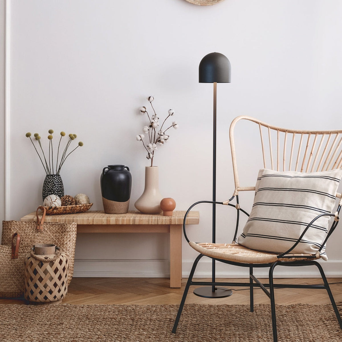 Rio 2 Light Dome Floor Lamp in matt black, slender stem and round base, shown beside a rattan chair and wooden bench in a living room setting.