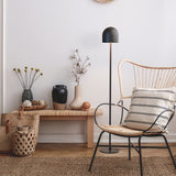 Rio 2 Light Dome Floor Lamp in matt black, slender stem and round base, shown beside a rattan chair and wooden bench in a living room setting.
