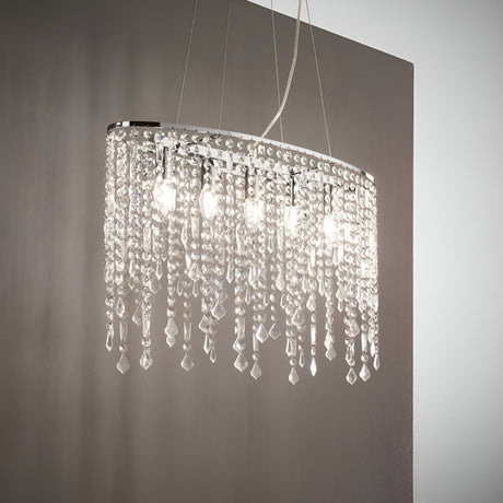 The Myst Crystal 5 Light Chandelier in Chrome, featuring an adjustable length and dangling crystal ornaments, hangs from the ceiling, illuminating the room with a bright glow. The elegant fixture is set against a neutral-colored wall, infusing the space with sophistication.