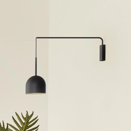 Rio Single Arm Dome Wall Light in matt black iron, with a slim right-angled arm from a rectangular wall plate and a cable-hung dome shade, shown against a plain wall near a potted plant.