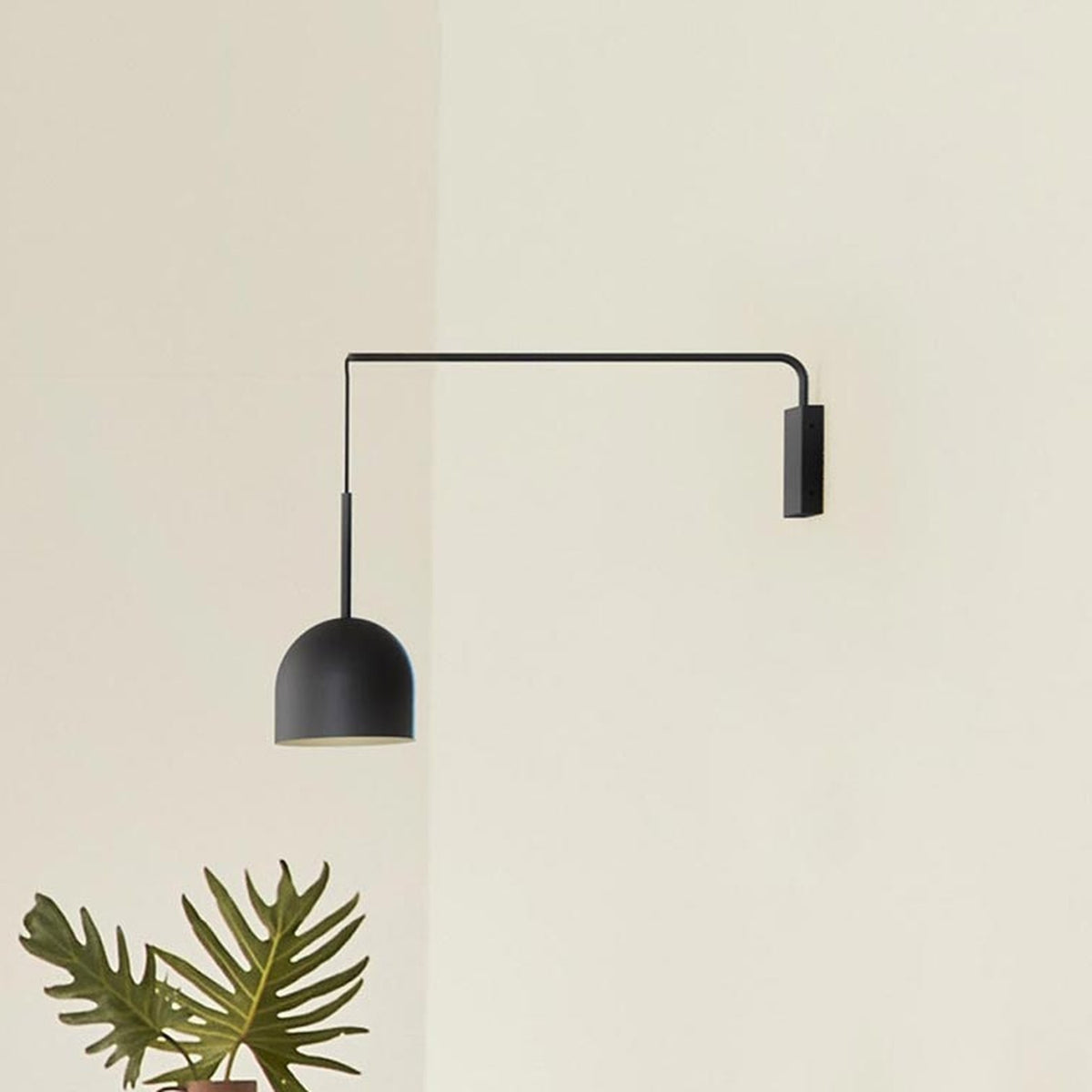 Rio Single Arm Dome Wall Light in matt black iron, with a slim horizontal arm and dangling cable leading to a rounded dome shade, shown against a plain wall with a plant below.