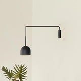 Rio Single Arm Dome Wall Light in matt black iron, with a slim horizontal arm and dangling cable leading to a rounded dome shade, shown against a plain wall with a plant below.