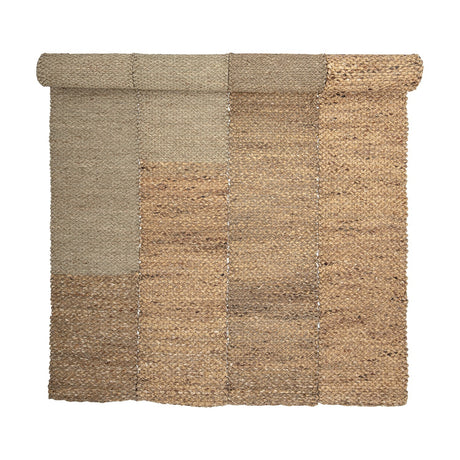 Enzov Seagrass Rug 245x150cm in Natural, partially rolled, showing four braided panels in varying earthy tones with visible stitched seams, on a plain white background.