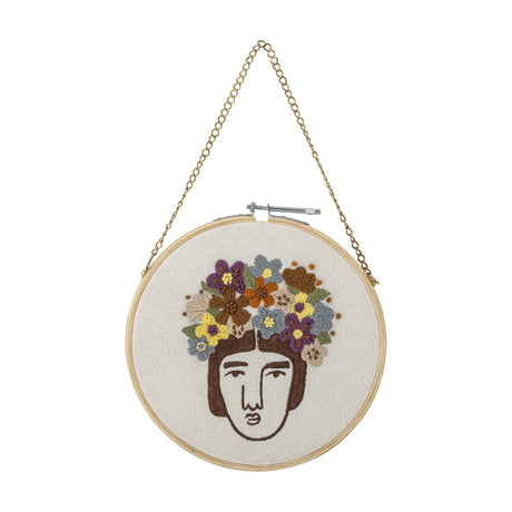 Round embroidered cotton hoop wall art featuring a line-drawn face with a textured multicoloured floral crown, shown against a plain white background.