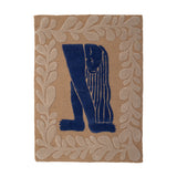 Atle Wool Wall Decor in Multi, a tufted wool hanging with a navy figure bent forward, hair falling in strands, framed by a tan leafy sprig border on a plain background.