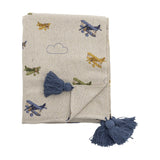 Plane Cotton Throw in beige, folded cotton blanket patterned with biplanes and outlined clouds, finished with chunky blue corner tassels, shown on a plain white background.