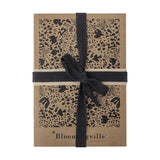 Anniversary Notebook in brown paper with a black bird, floral and leaf pattern, tied with a black woven ribbon, on a plain white background.