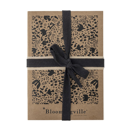 Anniversary Notebook in brown paper with a black bird, floral and leaf pattern, tied with a black woven ribbon, on a plain white background.