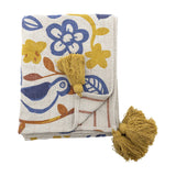Figuria Cotton Throw in multicoloured folded with mustard corner tassels, featuring printed flower, leaf and bird motifs in blue, yellow and rust on a cream background.