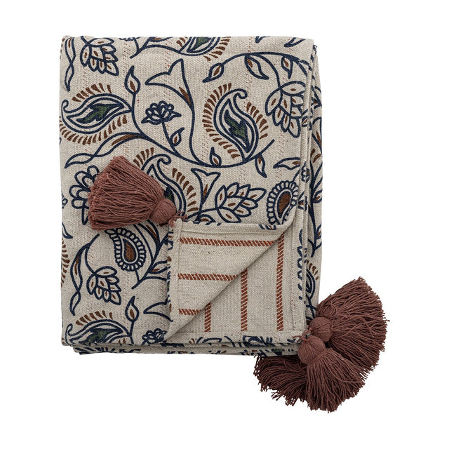 Folded cotton blend throw in cream with a navy paisley and floral print, striped reverse side, and rust-brown corner tassels, shown on a white background.
