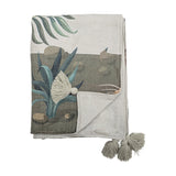 Peacock Throw in green, folded to show a printed garden scene with palm fronds, spiky leaves and stones, plus grey tassel trim, against a plain white background.