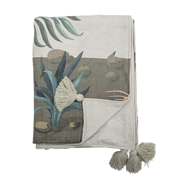 Peacock Throw in green, folded to show a printed garden scene with palm fronds, spiky leaves and stones, plus grey tassel trim, against a plain white background.