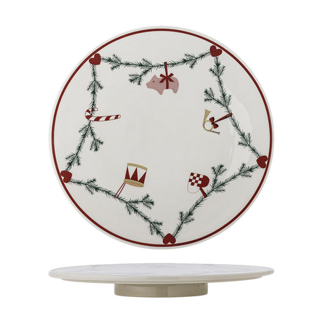 White stoneware lazy Susan with a fir garland and heart motifs linking a pig, candy cane, drum, horn and flag, on a low round base, plain white background.