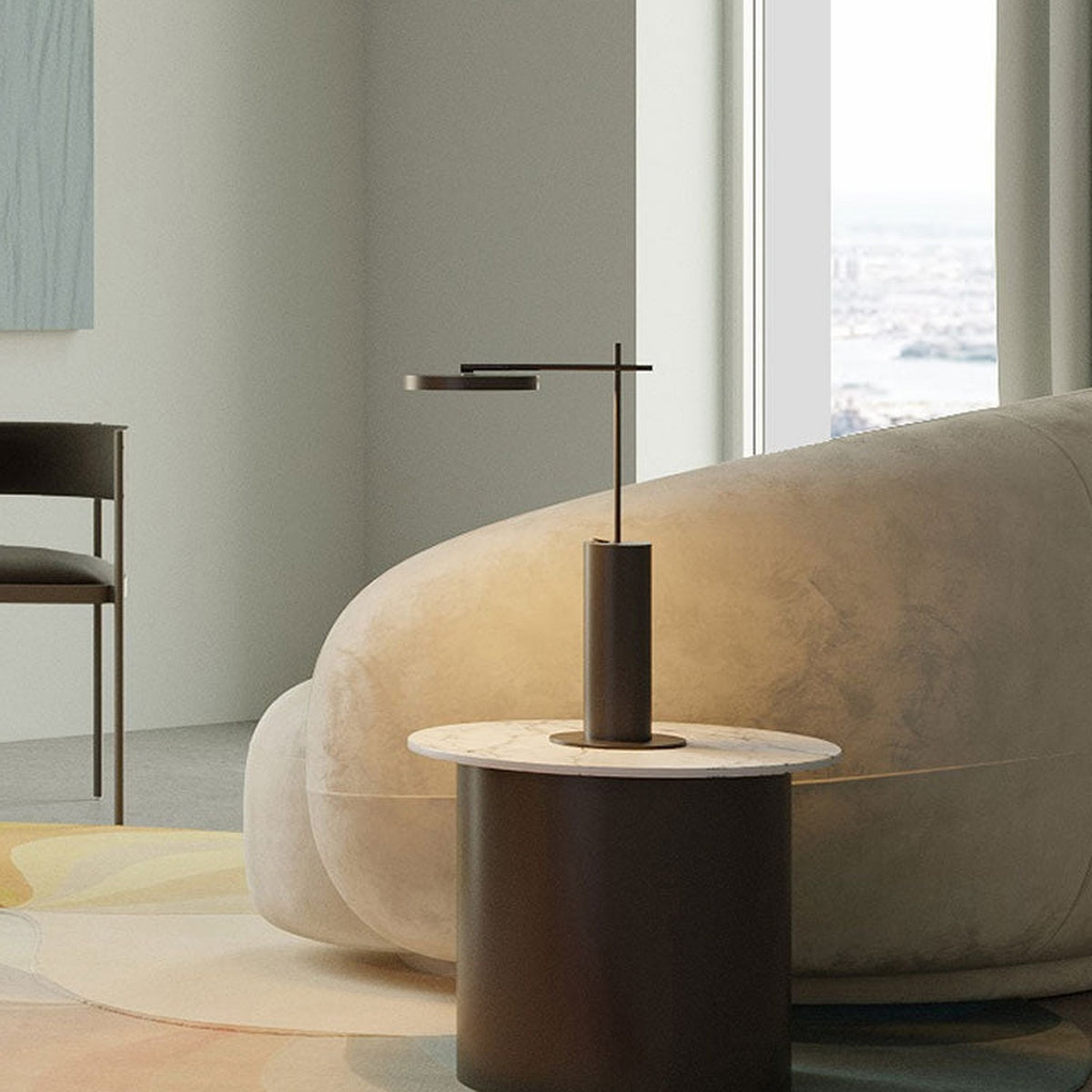Rafaela LED Disc Table Lamp in matt black with slim cylindrical base and flat disc head, shown lit on a marble side table beside a beige sofa.