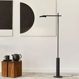 Rafaela LED Disc Floor Lamp in matt black, slim iron upright on a round base with a horizontal arm holding the aluminium disc head, shown in a living room beside abstract wall art.