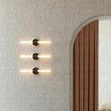 Three Roos LED Cylinder Disc wall lights in matt black, each with a round disc and horizontal light bar, mounted vertically on a plastered wall beside an arched wooden doorway.