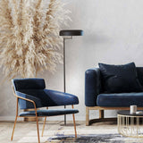Rea floor lamp with matt black dome shade on an adjustable swing arm and slim iron stem, shown beside a navy armchair and sofa in a living room setting.