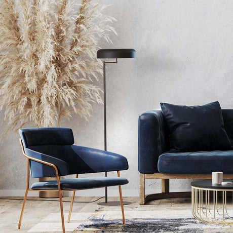 Rea floor lamp with matt black dome shade on an adjustable swing arm and slim iron stem, shown beside a navy armchair and sofa in a living room setting.