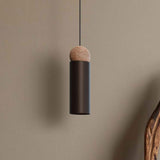 Rocio Wood Pendant Light in matt black, slim cylindrical metal shade topped with a round ash wood ball, hung on a textile cable against a plain neutral wall.