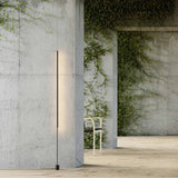 René LED Floor Lamp in matt black, a slim vertical light bar on a cylindrical base, shown glowing warm white against a concrete wall in a plant-filled interior.