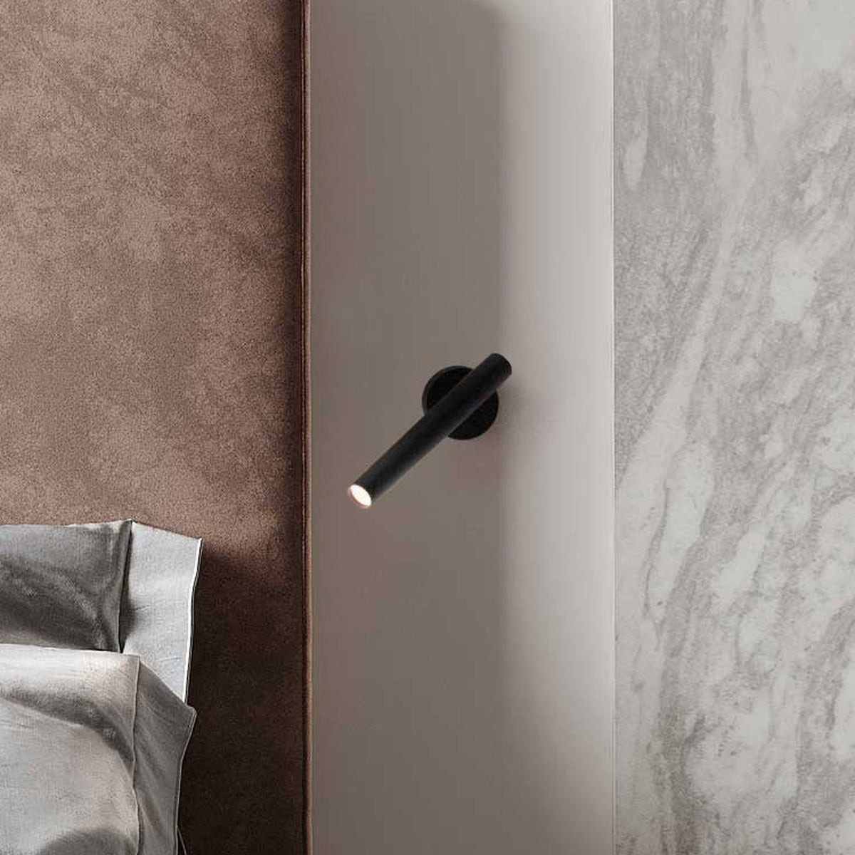 Rui LED Adjustable Wall Light with Reading Light in matt black, slim cylindrical tube on a short pivoting arm, mounted bedside above a bed with soft fabric headboard and marble wall panel.